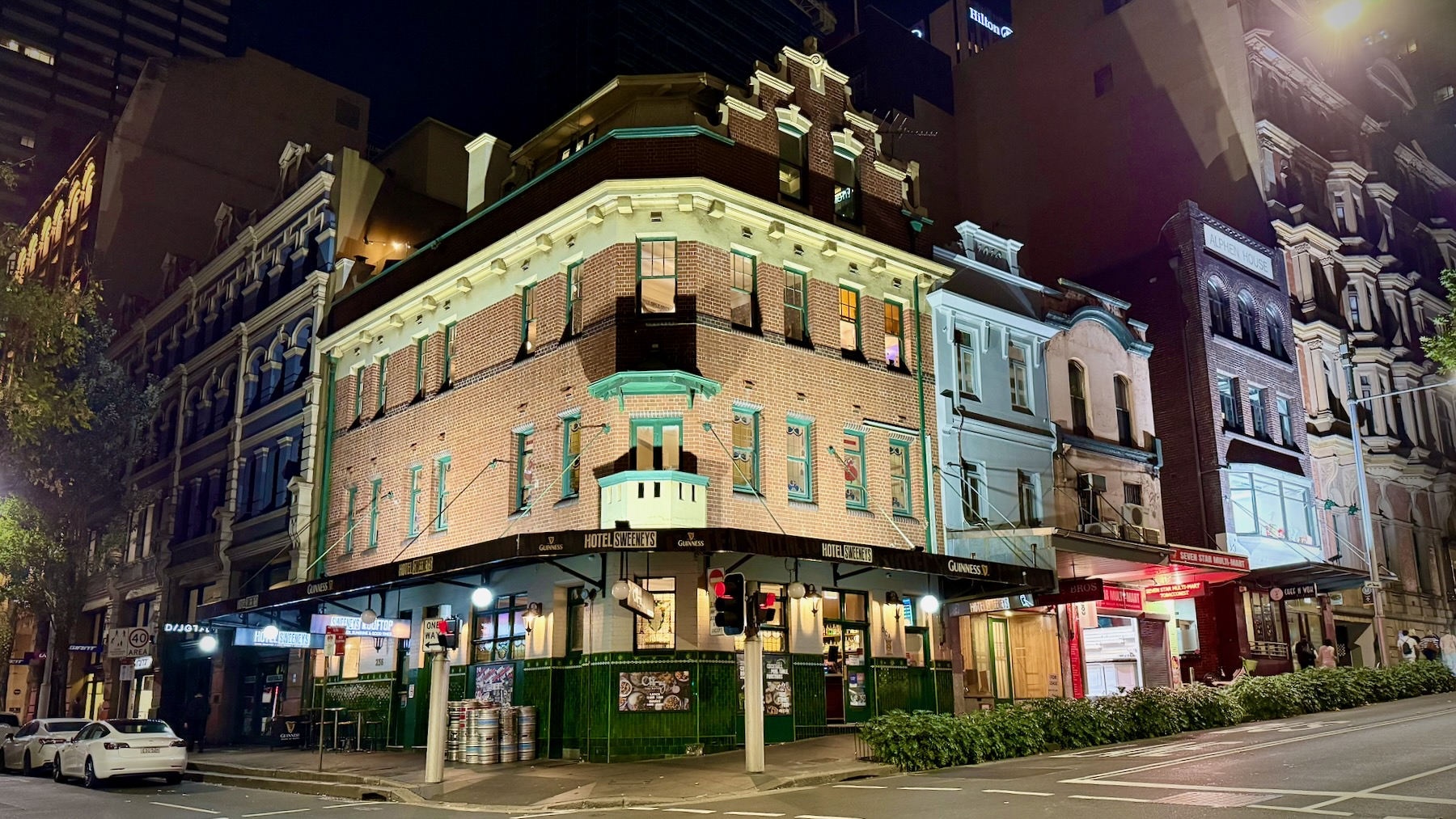 It’s nighttime. A typical old Sydney pub dominates the corner of two typical old Sydney city streets. It has four floors. The ground floor yellow-painted brick above deep green tiles. The next two floors up are reddish-brown brick. And up top there’s a brick level inset behind a terrace, with gable windows, Empty beer kegs are stacked by one door. The edge of the verandah is black, and is labelled “Hotel Sweeney’s” alternating with “Guinness”. Window trim and such are painted mid-green.