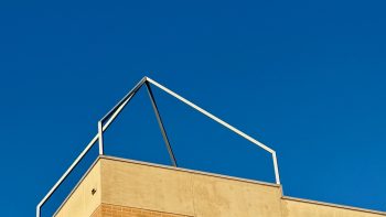Most of the image is a clear bright blue sky. At the bottom, the top of a cream brick building enters the frame, on top of which is the outline of a pyramid made of steel tubes. For some reason. Decoration I guess.