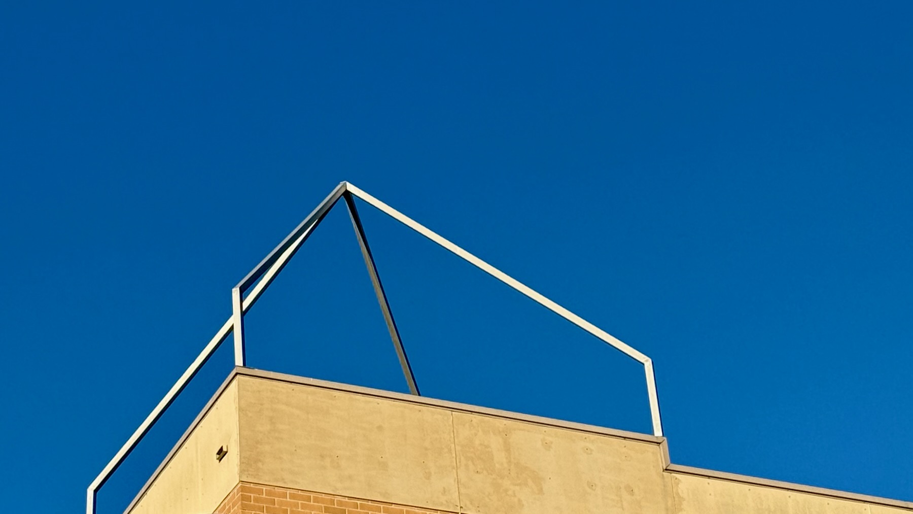 Most of the image is a clear bright blue sky. At the bottom, the top of a cream brick building enters the frame, on top of which is the outline of a pyramid made of steel tubes. For some reason. Decoration I guess.