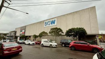 A view along a suburban street on a grey rainy day. On the other side of the rod, behind a line of cars waiting for the loghts to change is a big grey windowless concrete box, three storeys high. At the centre of the upper level is a big sign reading “BIG W” in blue on white. On the far left is signage reading “Campsie Centre”, “Tong Li Supermarket” and the same in Chinese, “Australia Post”, “Big W” again, and a blank red rectangle.
