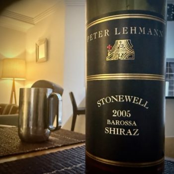 A bottle of wine with a black label sits on a table. The label reads: Peter Lehmann Stonewall 2005 Barossa Shiraz. In the background, out of focus, is a metal mug and, further back, lamps and such.
