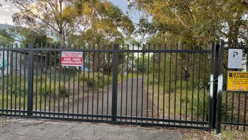 A black steel gate with spikes and locks and warning signs, behind which is a paved pathway along Cooks River.