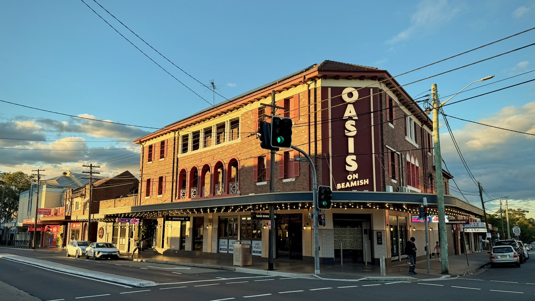 Looking across a suburban street to a three-storey red brick pub. The ground floor’s walls are tiled in yellow. The first floor has arched windows, while the second floor has square. Late afternoon sunlight strikes the left-hand side of the building. On the corner facing us, in the shade, is an illuminated sign reading “Oasis on Beamish”. The awning over the footpath is blue, its underside studded with glowing lightbulbs.