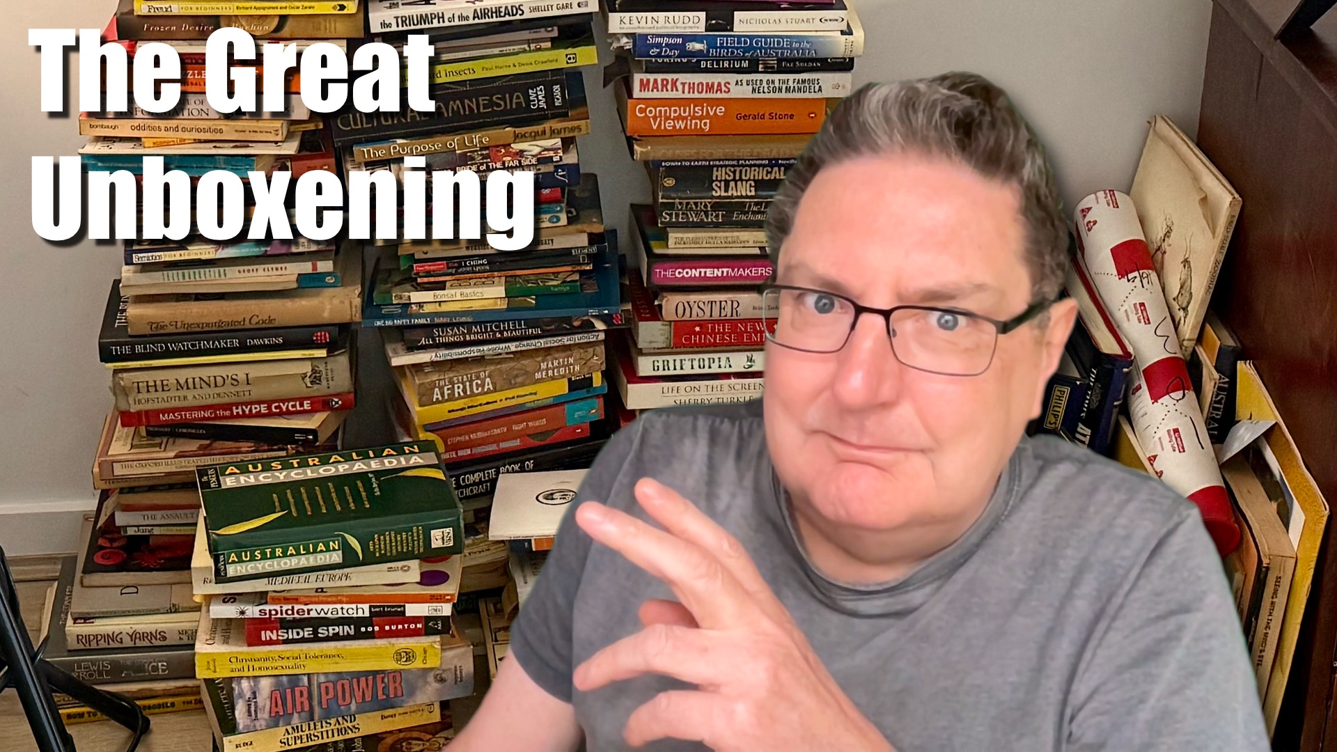 A clean-shaven white man with greying hair wears a grey t-shirt and spectacles. Yes, it’s Stilgherrian. He looks at the camera and gestures vaguely with his left hand. Behind him, on the floor, there’s a bunch of books stacked against the wall, untidily.