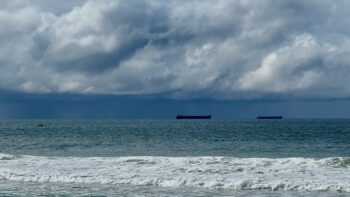 Looking out over the Pacific Ocean. On the right-hand side of the photo, riding the mackerel blue waters, are two big black bulk carrier ships, their red hulls high out of the water, their bows pointing to the right. On the far left, closer to us, is a small fishing boat. In the immediate foreground, waves break on the shore. Above are lumpy clouds, and in the in distance there is rain.
