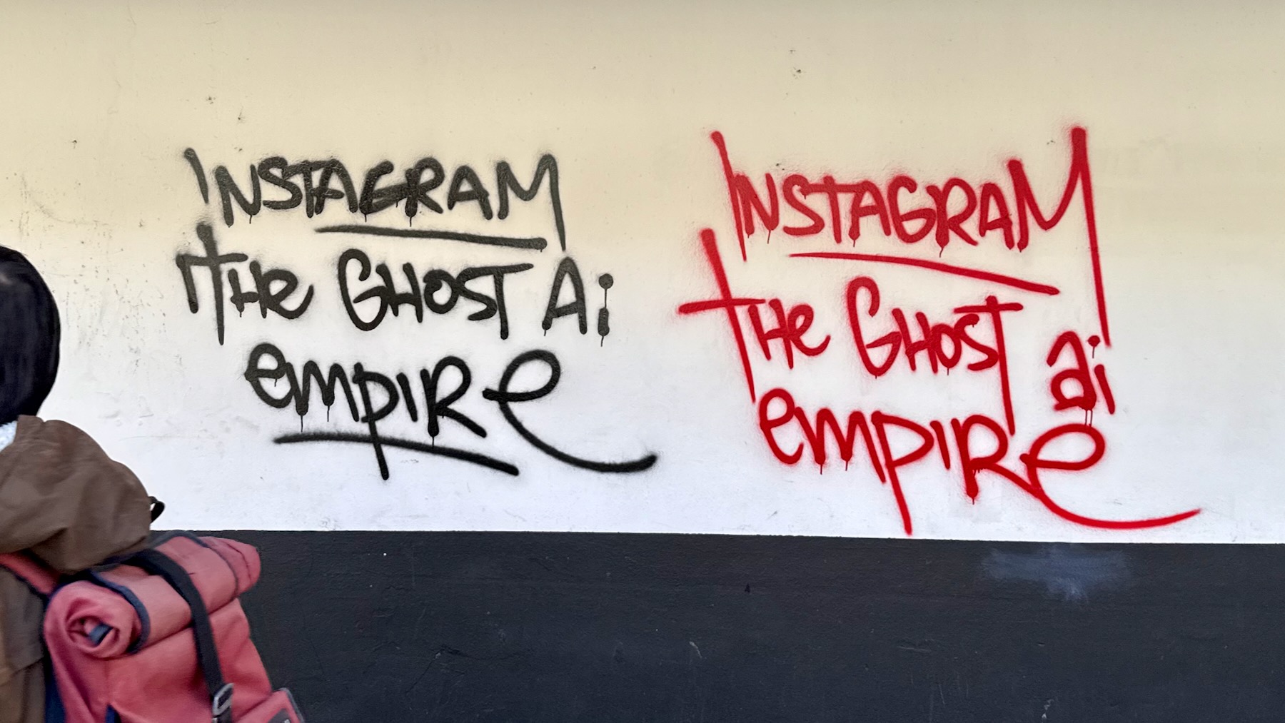 On a cream-coloured wall is scrawled in black the graffiti "Instagram the ghost AI empire", and the same again in red. On the left, a young man with a red backpack is disappearing out of frame.