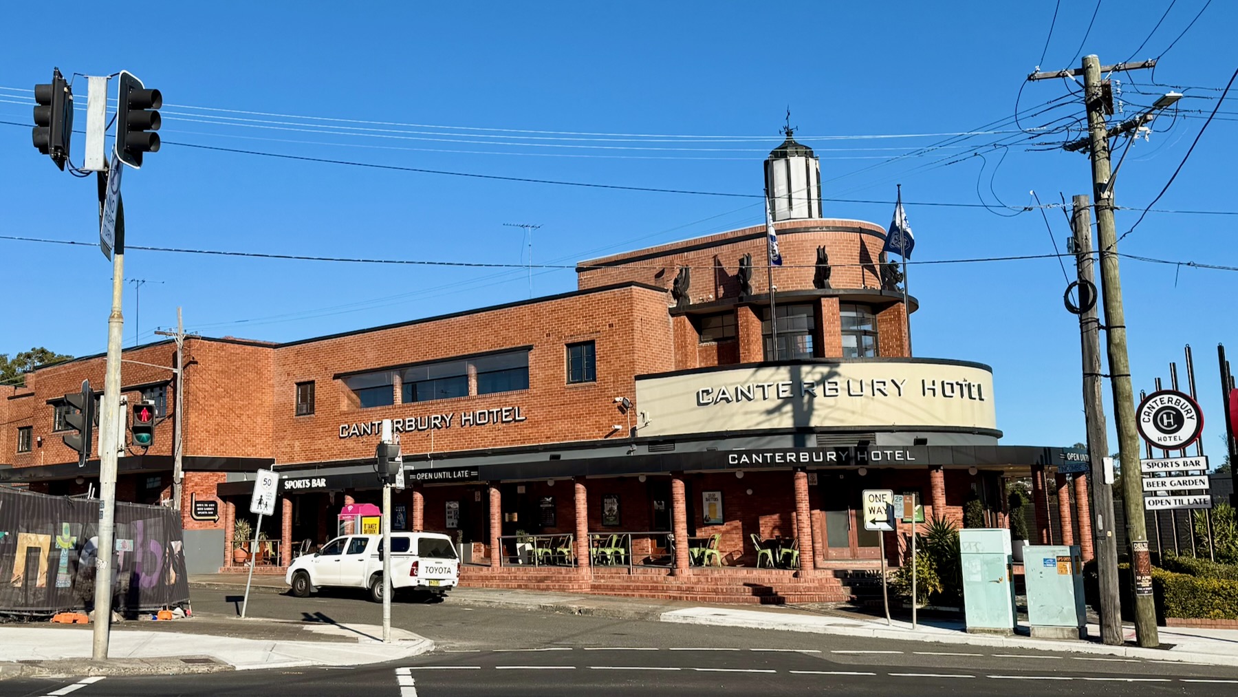 A red brick Australian suburban pub in the bright winter sunlight. On the left, the pub’s front, it’s a semi-circular arrangement with a balcony topped by a series of sculpted pegasuses, and right at the top a lighthouse-like structure — which once was an actual lighthouse! It’s on the other side of the highway from us. On its left is a tall mast for traffic lights. On the right, a pair of wooden utility poles for electricity, telephone, and internet.