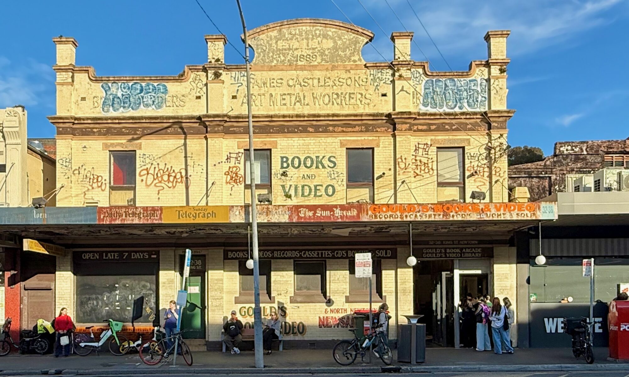 A two-storey Victorian commercial building glows in the late afternoon light. It’s painted a faded creamy yellow. The upper level, with an ornate facade, reads “Books and Videos” in somewhat faded text, and in even older text “1889 James Castle & Sons Art Metal Workers”. There’s more text, but it’s obscured by graffiti. The awning has faded signage for the Daily Telegraph, Sunday Telegraph, The Sun-Herald, and, finally, “Gould’s Books & Video, Records & Cassettes Bought & Sold”. At street level the Gould’s signage is repeated. Bicycles are locked to some parking signs. And on the right of the building there’s a door into which winds a queue of young people waiting to get in.