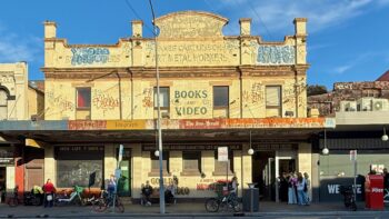 A two-storey Victorian commercial building glows in the late afternoon light. It’s painted a faded creamy yellow. The upper level, with an ornate facade, reads “Books and Videos” in somewhat faded text, and in even older text “1889 James Castle & Sons Art Metal Workers”. There’s more text, but it’s obscured by graffiti. The awning has faded signage for the Daily Telegraph, Sunday Telegraph, The Sun-Herald, and, finally, “Gould’s Books & Video, Records & Cassettes Bought & Sold”. At street level the Gould’s signage is repeated. Bicycles are locked to some parking signs. And on the right of the building there’s a door into which winds a queue of young people waiting to get in.