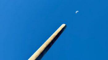 A bright blue cloudless sky. A long tube — is it metal? is it concrete? — extends from the lower left and thrusts upwards to the right, where it points directly at the half-full Moon. Yes, it does look like a giant Anti-Moon Cannon, but it’s really a ventilation device of some sort.