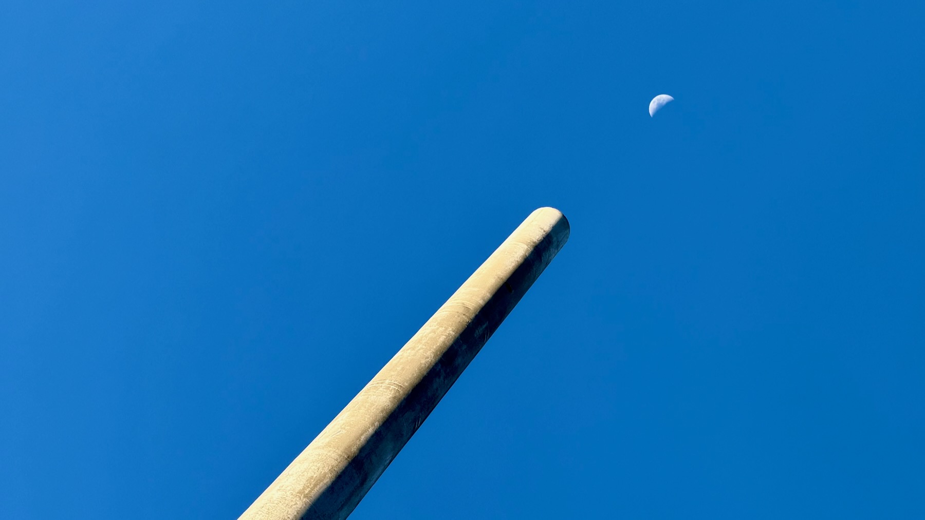 A bright blue cloudless sky. A long tube — is it metal? is it concrete? — extends from the lower left and thrusts upwards to the right, where it points directly at the half-full Moon. Yes, it does look like a giant Anti-Moon Cannon, but it’s really a ventilation device of some sort.