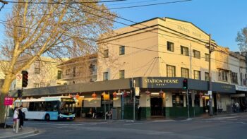 A street corner in suburban Sydney. We’re looking diagonally across the intersection to an inter-war commercial building, probably 1930s by my guess, which has been converted into a pub. The ground level is painted cream with a lower section of dark green tiles. Above are two floors of cream-painted walls, topped with the legend “Station House”. The awning also reads “Station House Hotel”, and is hung with red Chinese lanterns. Either side of the image are London plane tree, bare of leaves because it’s only the very start of spring. A blue and white bus is pulling up from the left.