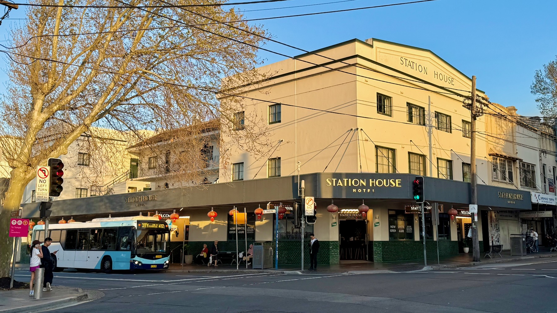 A street corner in suburban Sydney. We’re looking diagonally across the intersection to an inter-war commercial building, probably 1930s by my guess, which has been converted into a pub. The ground level is painted cream with a lower section of dark green tiles. Above are two floors of cream-painted walls, topped with the legend “Station House”. The awning also reads “Station House Hotel”, and is hung with red Chinese lanterns. Either side of the image are London plane tree, bare of leaves because it’s only the very start of spring. A blue and white bus is pulling up from the left.