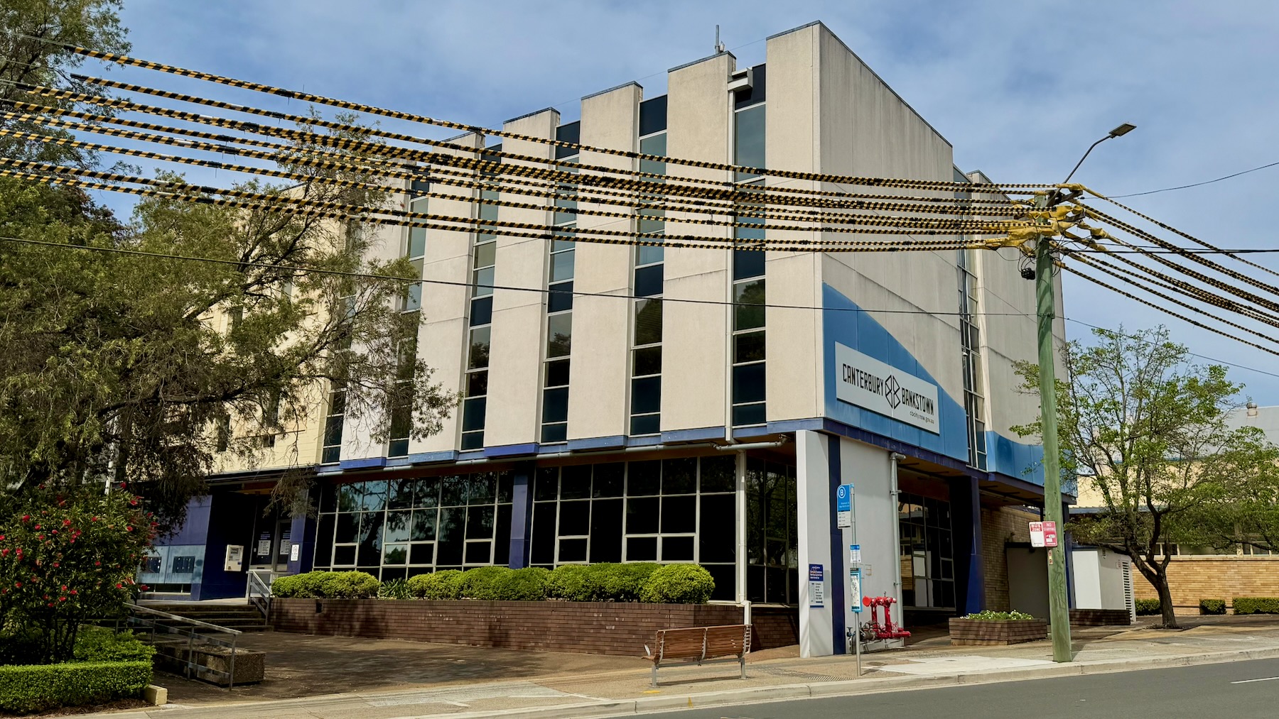 It’s a partly cloudy day. We’re looking at a modernist office building, three storeys high. It’s mostly grey concrete, with alternating vertical concrete and glass panels. The centre section has pale yellow panels with rectangular geometric designs. On the street side is the logo of the City of Canterbury-Bankstown. On the left are the trees of Lofts Garden, and there’s another tree on the right. But most importantly, all the electricity wires in front of the building have been fitted with black and yellow warning covers.